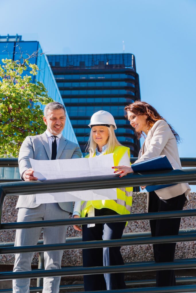 smiling architects and engineers reviewing blueprint, standing near contemporary office structure, collaborating on urban development project